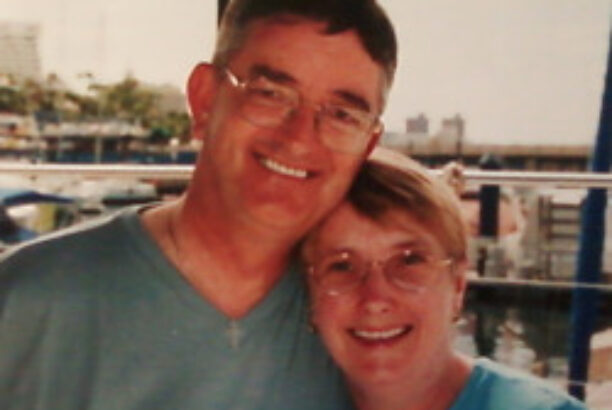 Husband and wife in their late 50s with arms around each other smiling at the camera