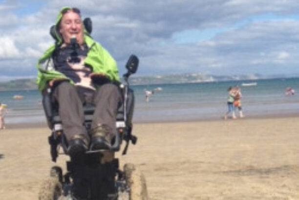Man in an electric wheelchair on the beach with his daughter buried in the sand in the foreground, both happy and smiling
